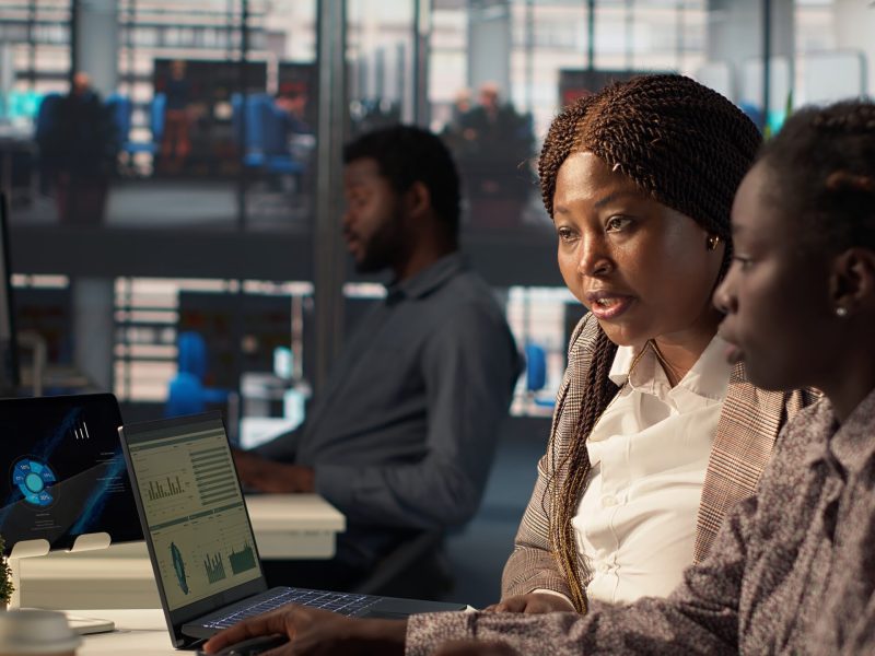 General manager reviews financial forecasts with an employee at desk, leading a session on project development and corporate goals. Collaborating to meet business objectives. Camera A.